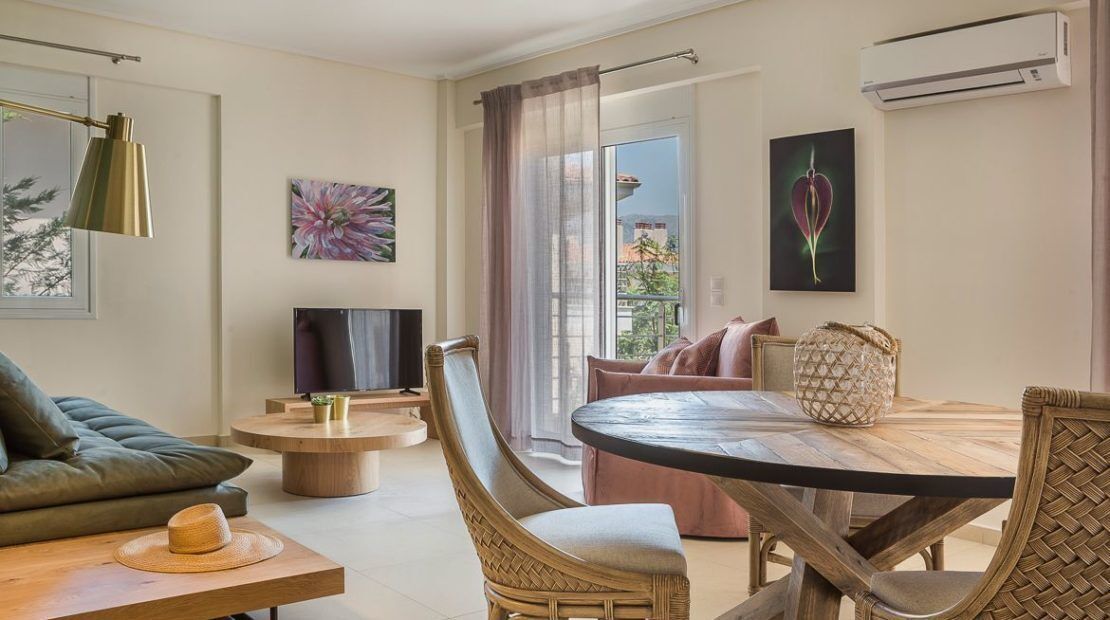 A modern living room with a sofa, a round wooden table, and a TV. There is a balcony with a view of buildings and trees.