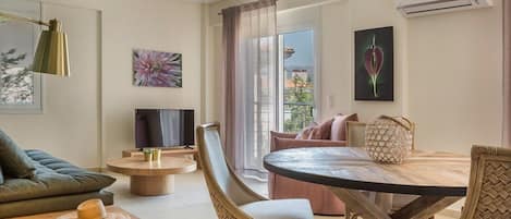 A modern living room with a sofa, a round wooden table, and a TV. There is a balcony with a view of buildings and trees.