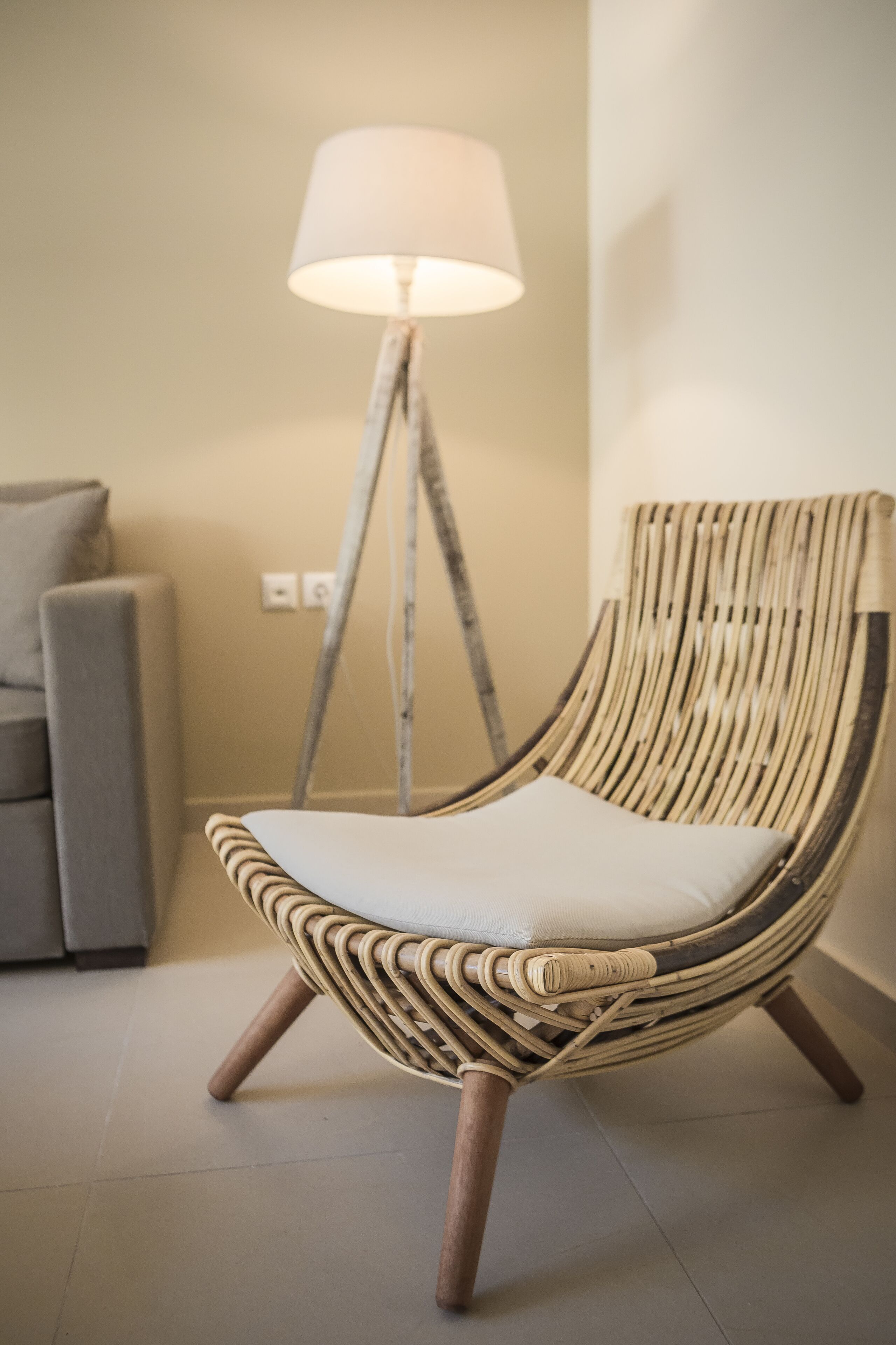 A wicker chair with a cushion, a floor lamp, and a grey sofa in a room with a tiled floor.