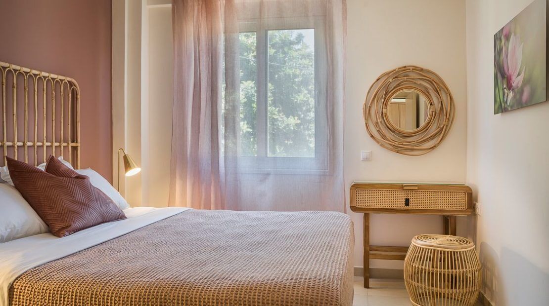 Une chambre à coucher avec un lit, une lampe de chevet, une fenêtre avec des rideaux, un miroir rond et une table d’appoint en osier.