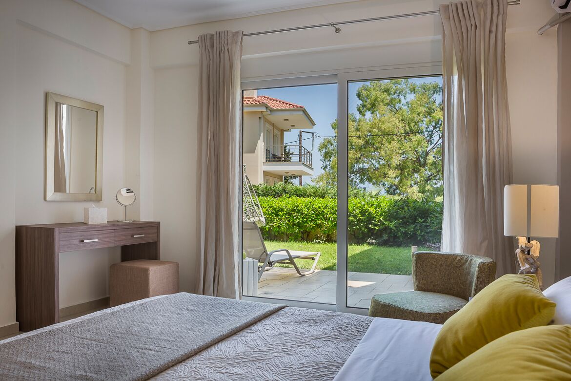 A hotel room with a bed, a desk, a chair, and a balcony with a view of a building and greenery.