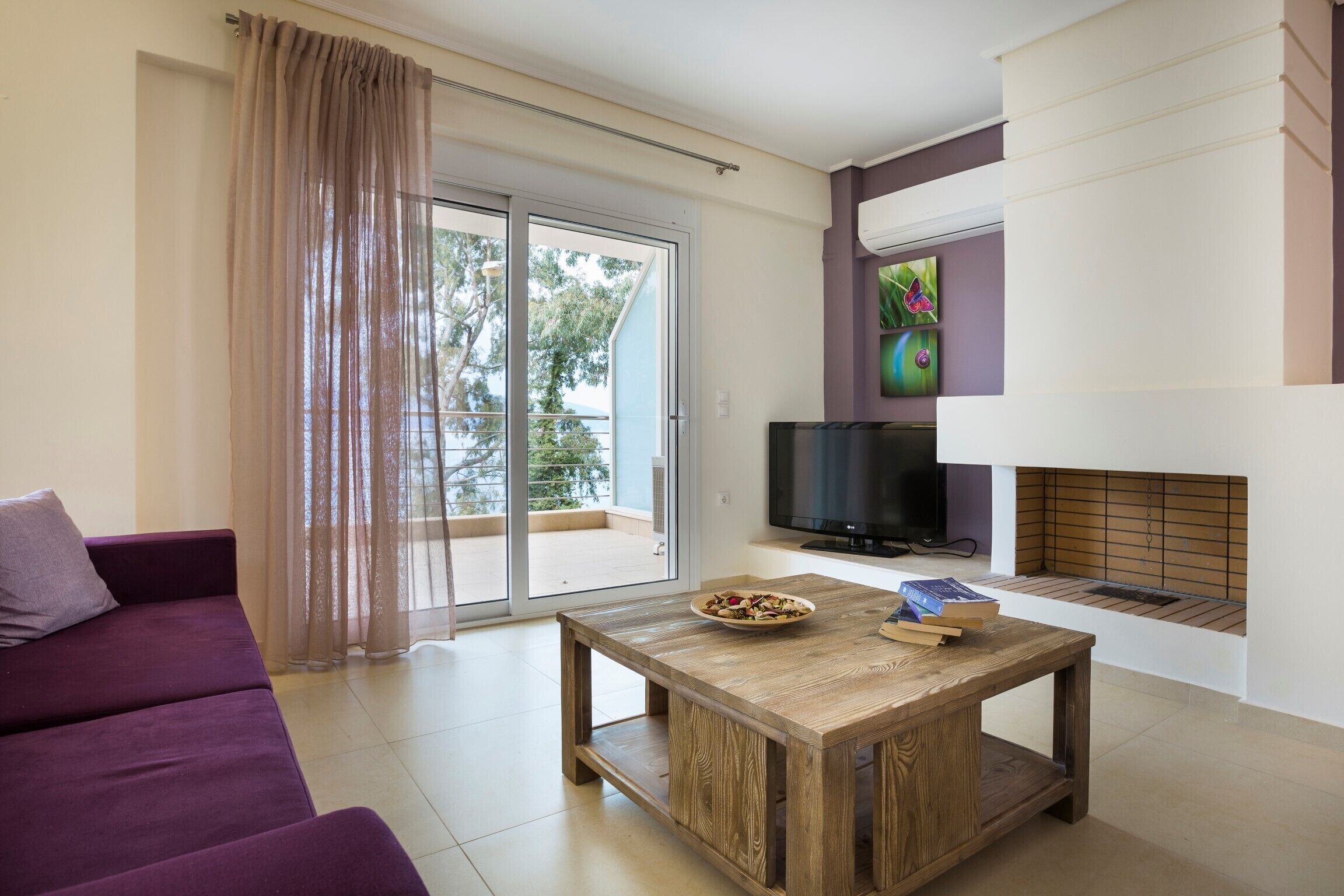 Ein modernes Wohnzimmer mit einem violetten Sofa, einem Couchtisch mit Büchern, einem Flachbildfernseher und einem Kamin.
