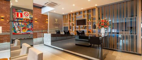 A modern hotel lobby with seating, a reception desk, and a large TV screen.