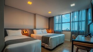 Habitación de hotel con dos camas, un escritorio y vistas de la ciudad a través de amplias ventanas.