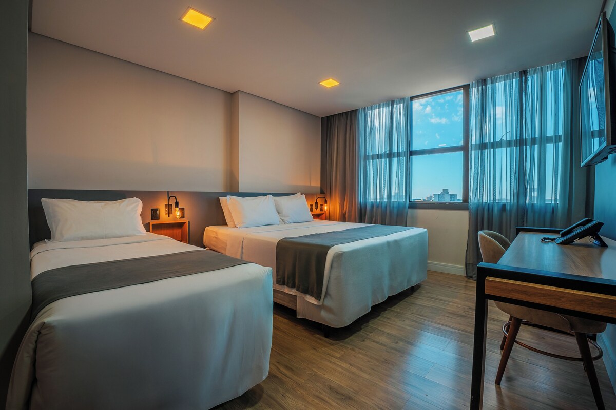 A hotel room with two beds, a desk, and a view of the city through large windows.