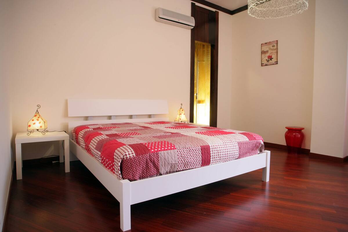 A bedroom with a white bed, a bedside table with a lamp, a red vase, and a wall-mounted air conditioner.