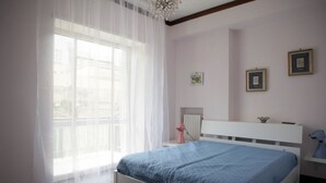 Ein Schlafzimmer mit Bett, einem Fenster mit durchsichtigen Vorhängen, einem Nachttisch mit Lampe und gerahmten Bildern an der Wand.