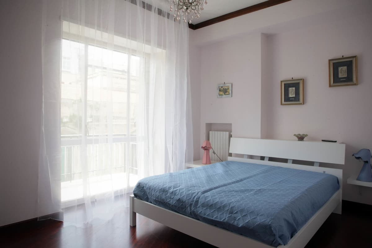 A bedroom with a bed, a window with sheer curtains, a bedside table with a lamp, and framed pictures on the wall.