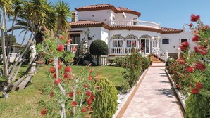 A large, white building with a red-tiled roof, surrounded by a well-maintained garden with various plants and a paved walkway leading to the entrance.