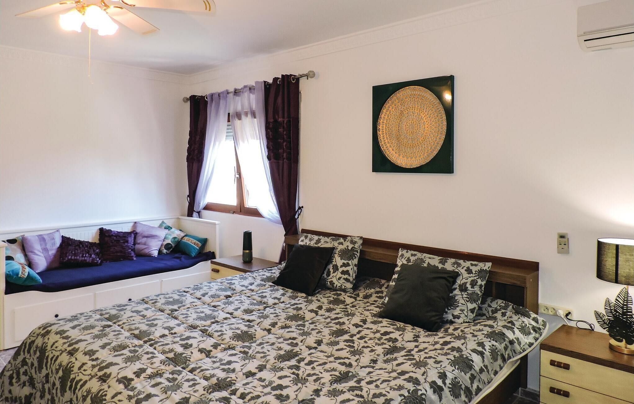 Ein Schlafzimmer mit Bett, einem Fenster mit Vorhängen, einem Deckenventilator, einer Wandklimaanlage und einer Wanddekoration.