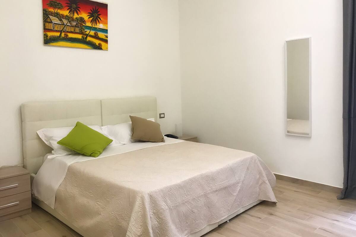 Una cama bien hecha con un cabecero color beige y almohadas, una mesita de noche pequeña, un espejo montado en la pared y un cuadro de una escena tropical.