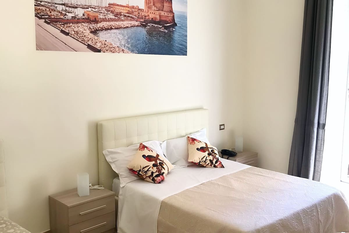 Habitación de hotel con cama, mesitas de noche, cortinas en la ventana y un cuadro colgado en la pared que representa una escena costera.