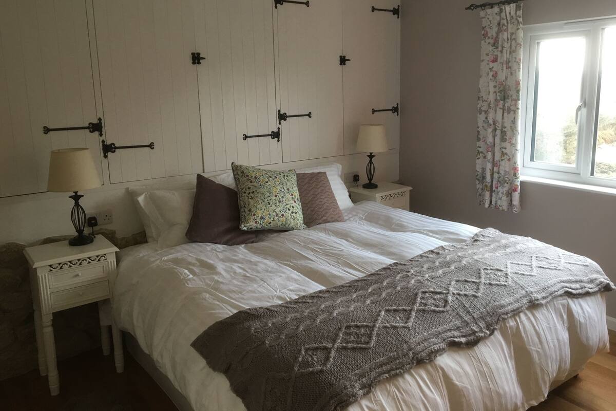A bedroom with a large bed, two bedside tables with lamps, a window with floral curtains, and a wall with decorative brackets.