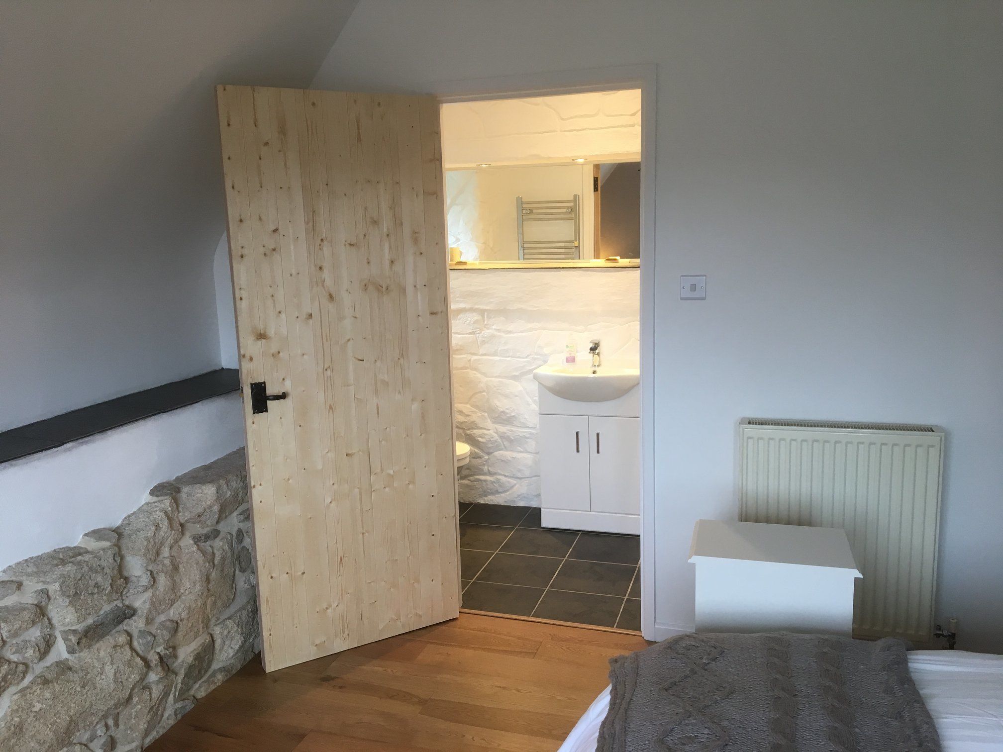 A bedroom with a wooden door, a stone wall, a bed with a blanket, a white bedside table, and a bathroom with a sink and cabinets.
