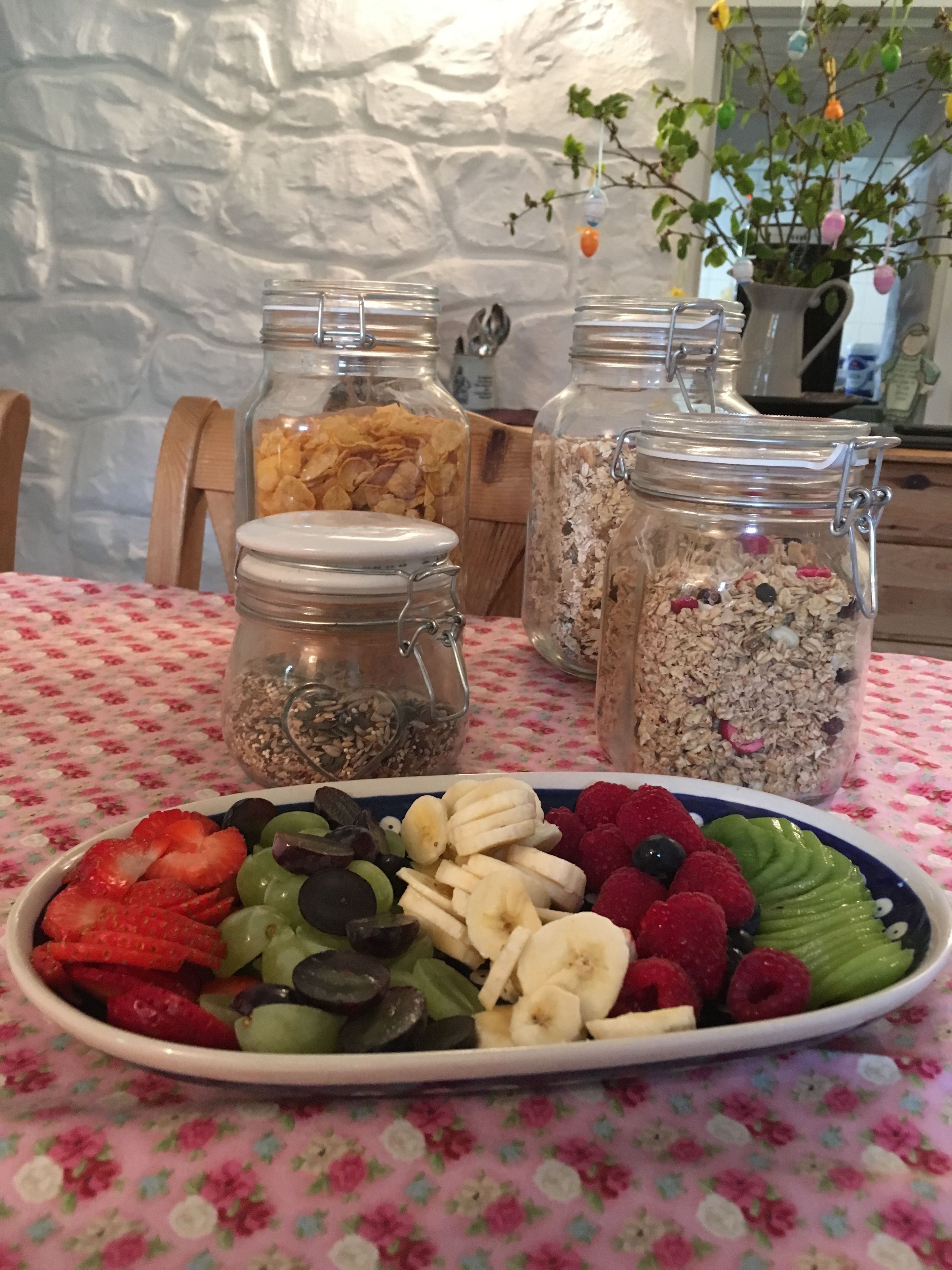 Een tafel met een tafelkleed met bloemen, een bord vol gemengd fruit en verschillende glazen potten met uiteenlopende inhoud.