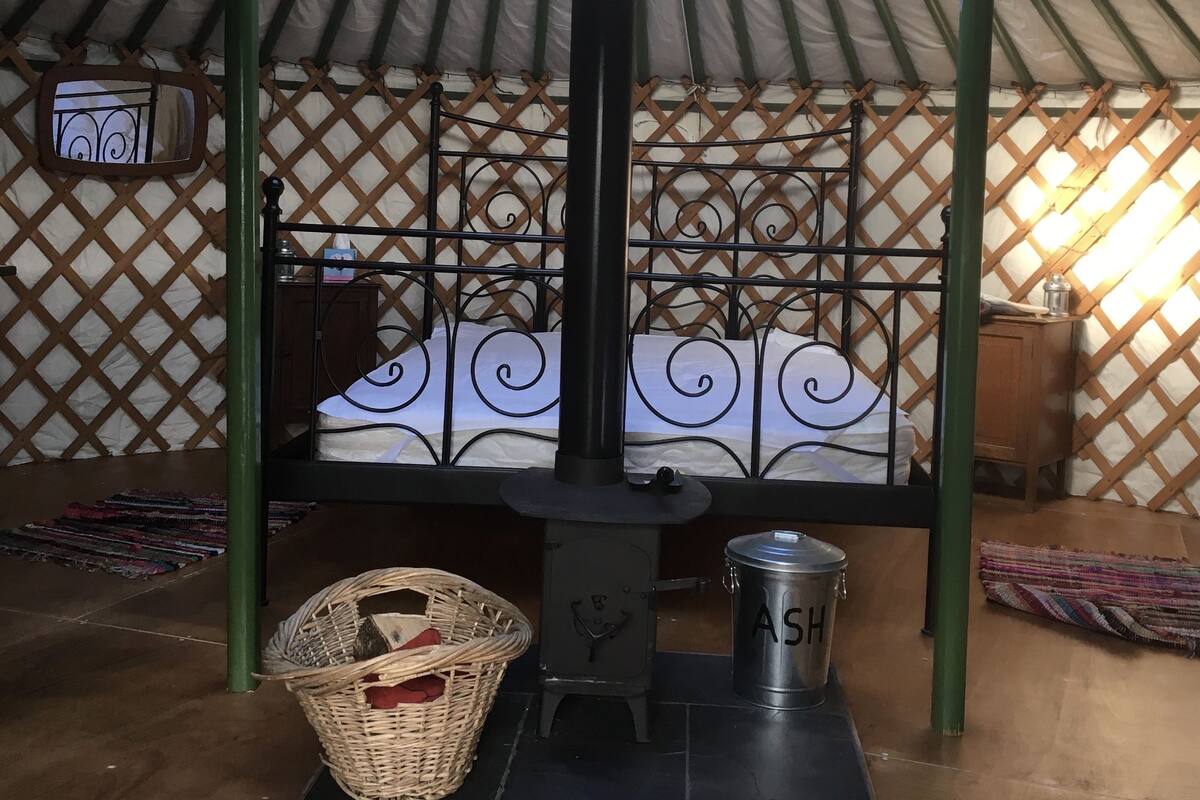 A traditional yurt interior with a bed, a metal stove, a woven basket, and a metal container labeled "ASH".