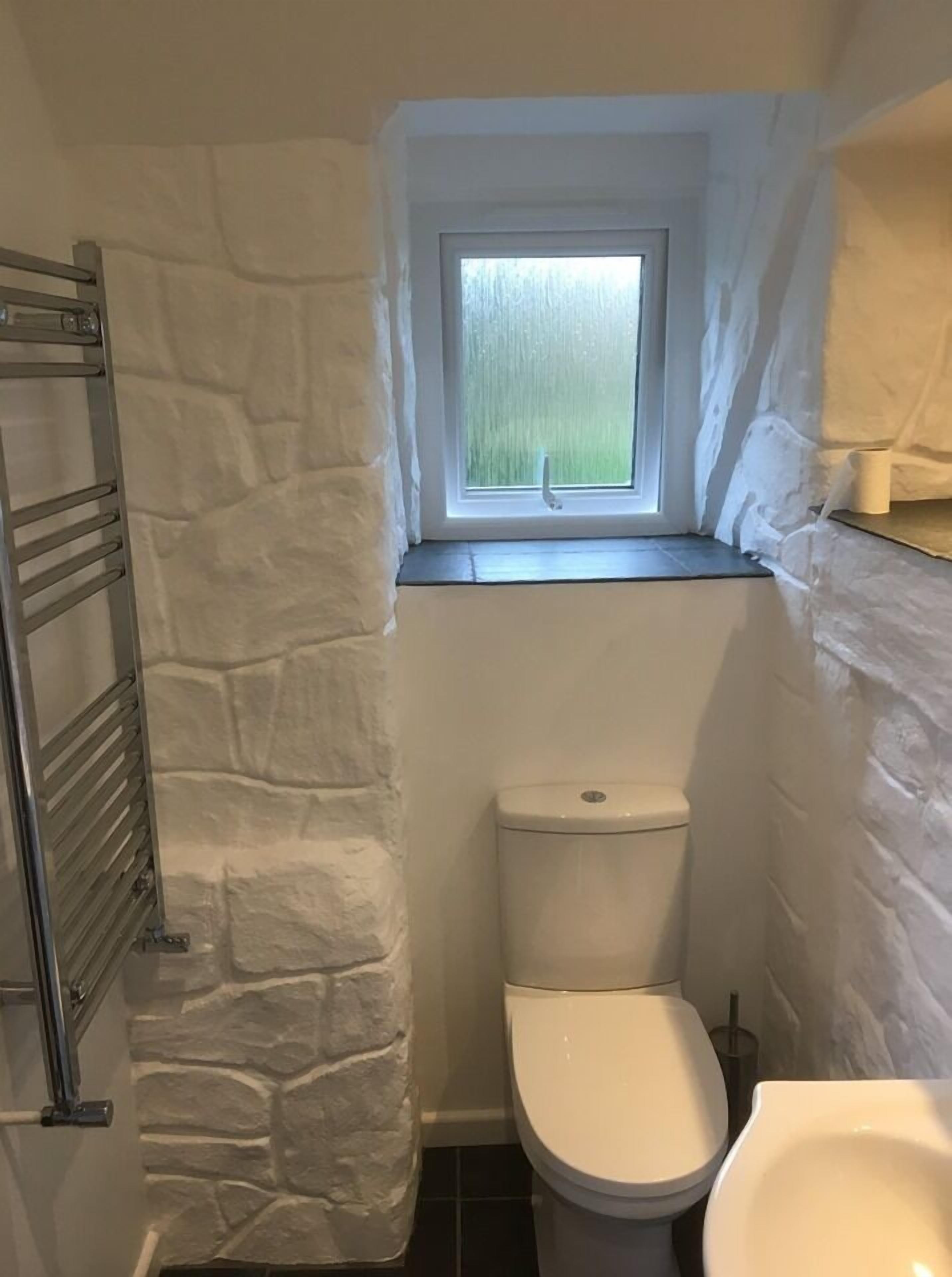 A small bathroom with a stone wall, a window, a toilet, and a metal towel rack.