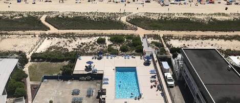 Zona de piscina en la azotea con sillones de descanso y sombrillas, con vista a una playa con sombrillas y gente.