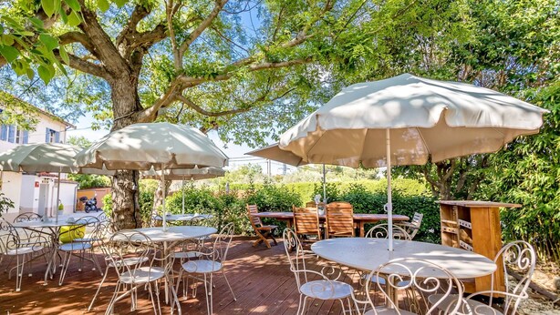 An outdoor patio with round tables and chairs, covered by large umbrellas, surrounded by greenery and trees. - Hôtel Des Moulins (Allauch)