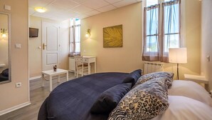A bedroom with a large bed, a desk, and a chair. There is a window with curtains, a radiator, and a wall-mounted lamp. - Hôtel Des Moulins (Allauch)