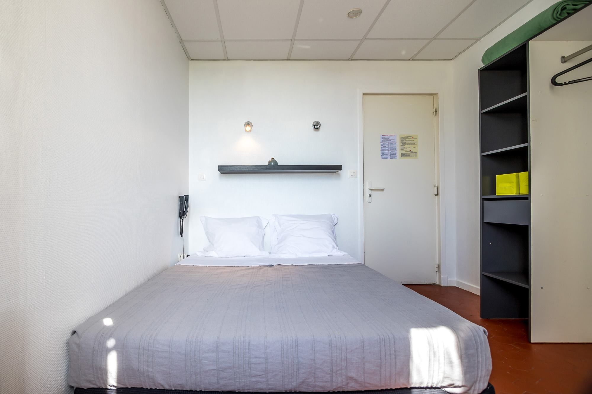 A single bed with a white bedspread in a room with white walls, a door, and a black shelving unit with a hanging garment.