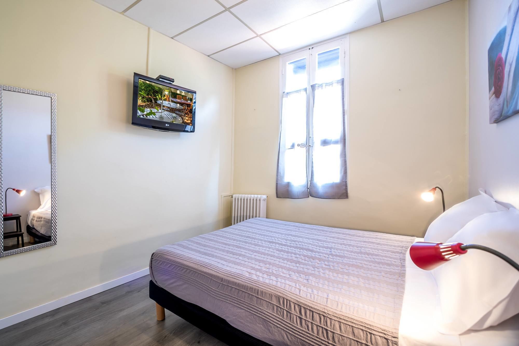 A bedroom with a bed, a TV mounted on the wall, a window with curtains, a radiator, and a bedside lamp.