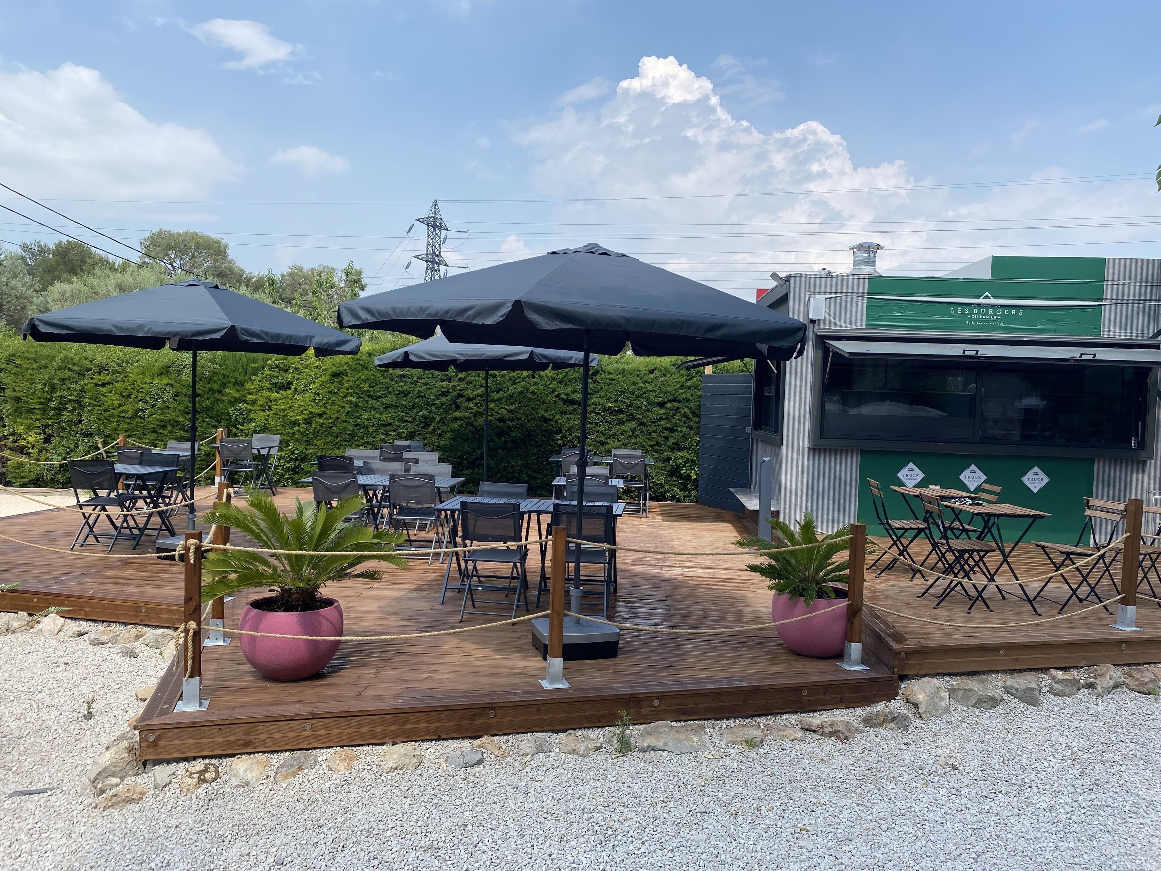 Une terrasse extérieure avec des tables et des chaises, entourée de verdure et bordée d’un bâtiment sur lequel on peut lire l’inscription « LES BURGERS ».