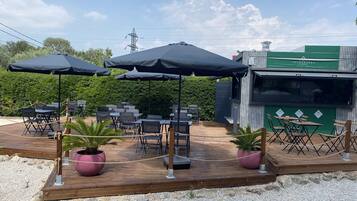 Une terrasse extérieure avec des tables et des chaises, entourée de verdure et bordée d’un bâtiment sur lequel on peut lire l’inscription « LES BURGERS ».