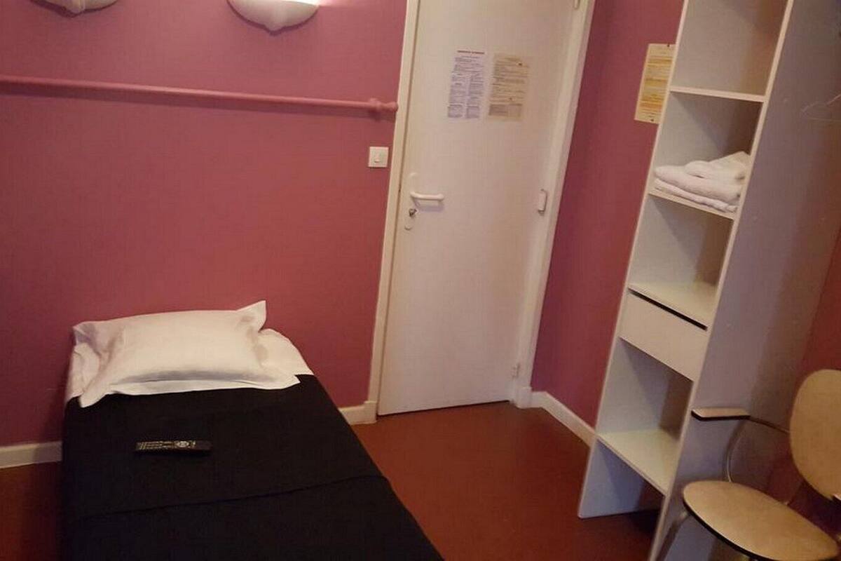 Een kleine, smalle kamer met een enkel bed, een witte deur en een plank met opgevouwen handdoeken.