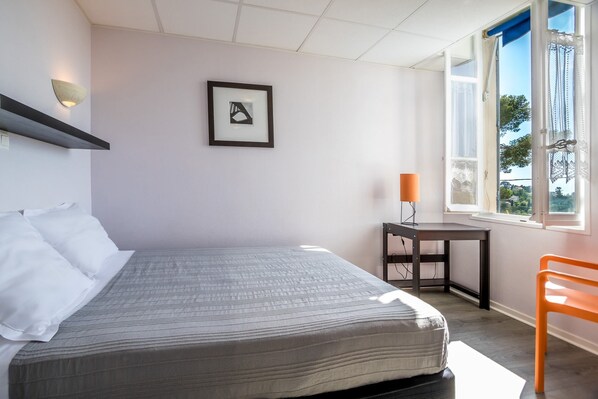 Une chambre à coucher comprenant un lit, un bureau, une chaise, une fenêtre et un luminaire fixé au mur.