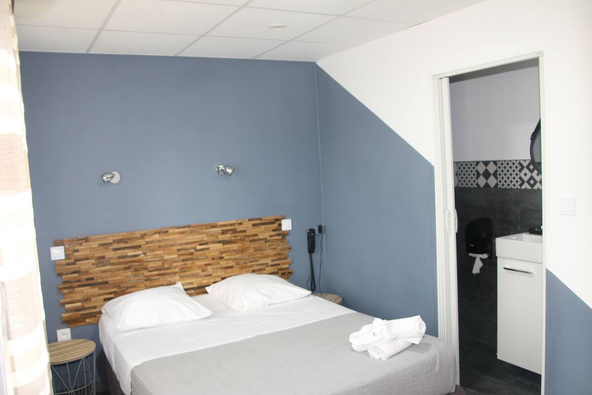Een hotelkamer met een houten hoofdbord, een bed met witte lakens, een klein rond krukje en een zichtbare badkamer door een geopende deur.