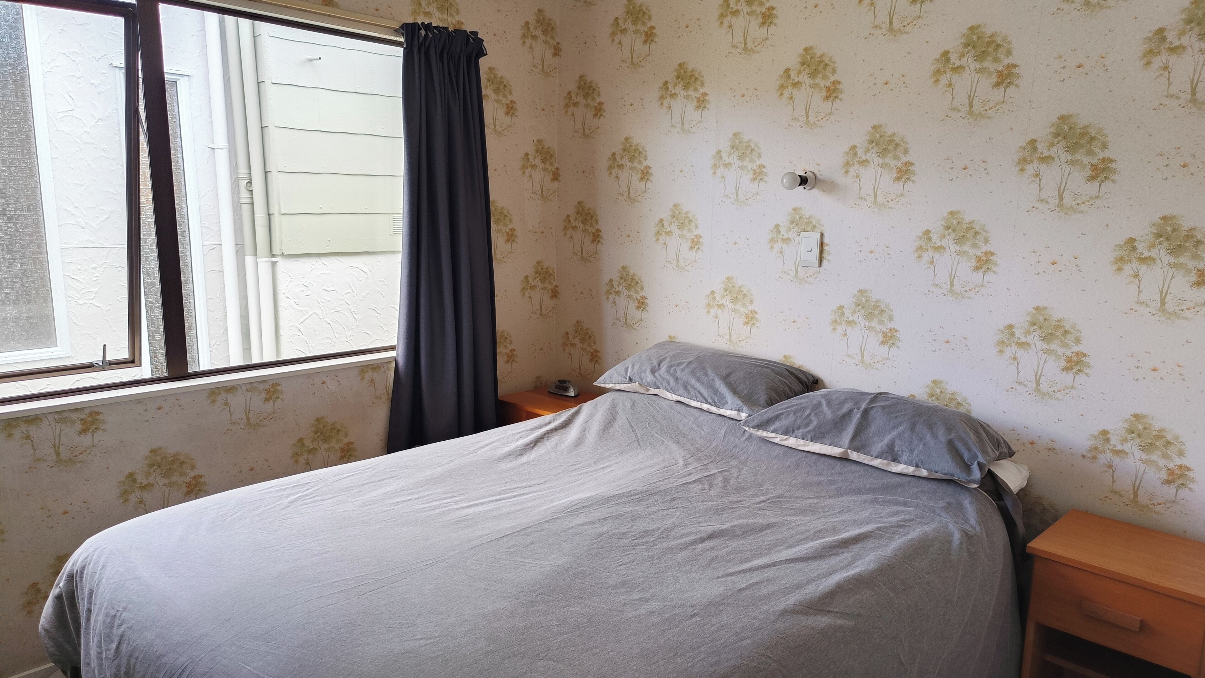 Ein Schlafzimmer mit einem Bett, einem Nachttisch, einem Fenster mit Vorhängen und einer Wand mit Baum-Motiv.