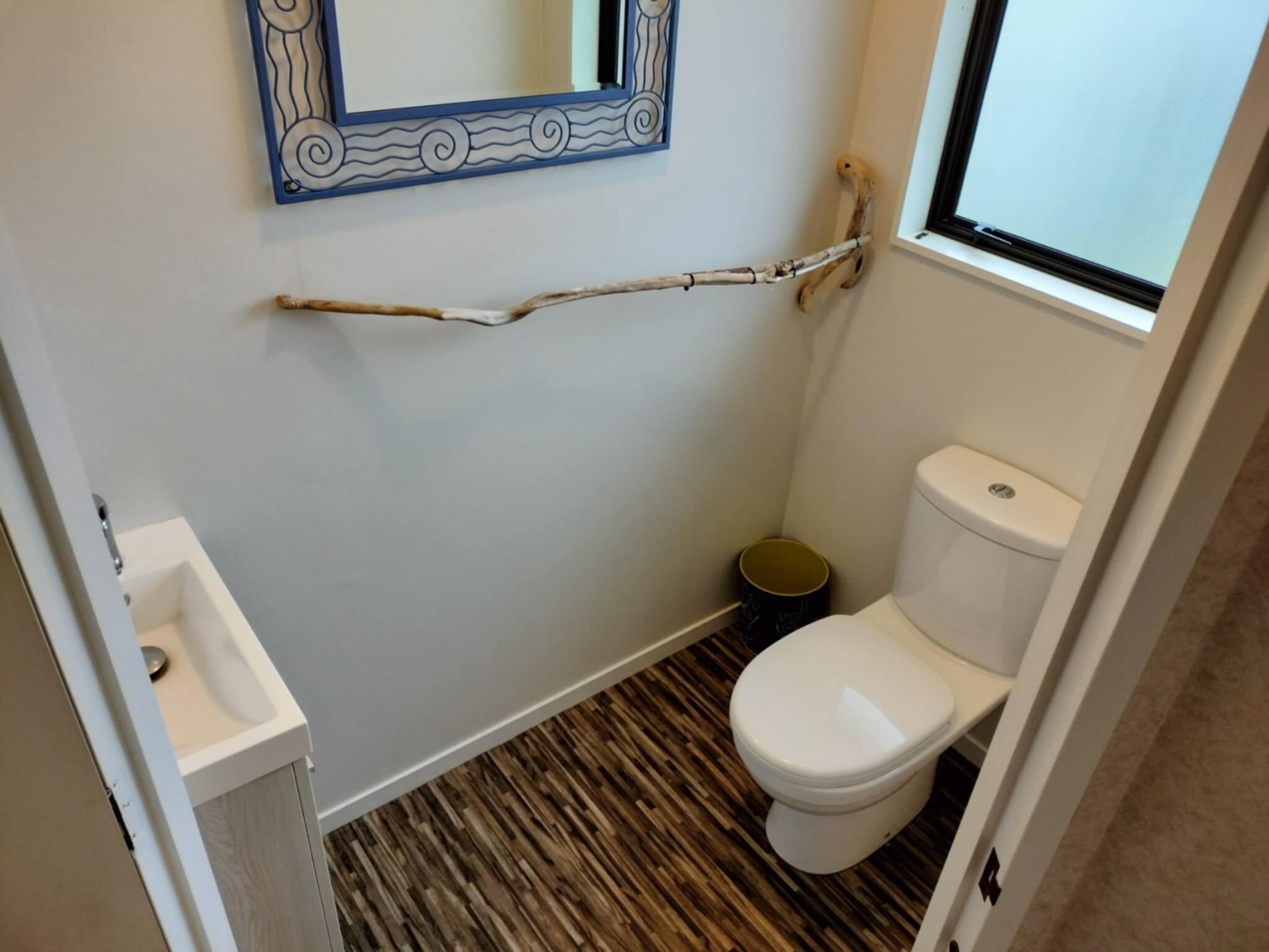 Un baño pequeño con inodoro blanco, piso de madera, lavamanos pequeño y espejo con marco decorativo.