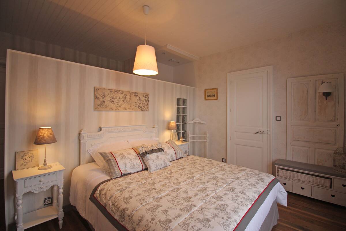 Un dormitorio con cama, mesitas de noche, lámpara colgante, armario y un cuadro en la pared.