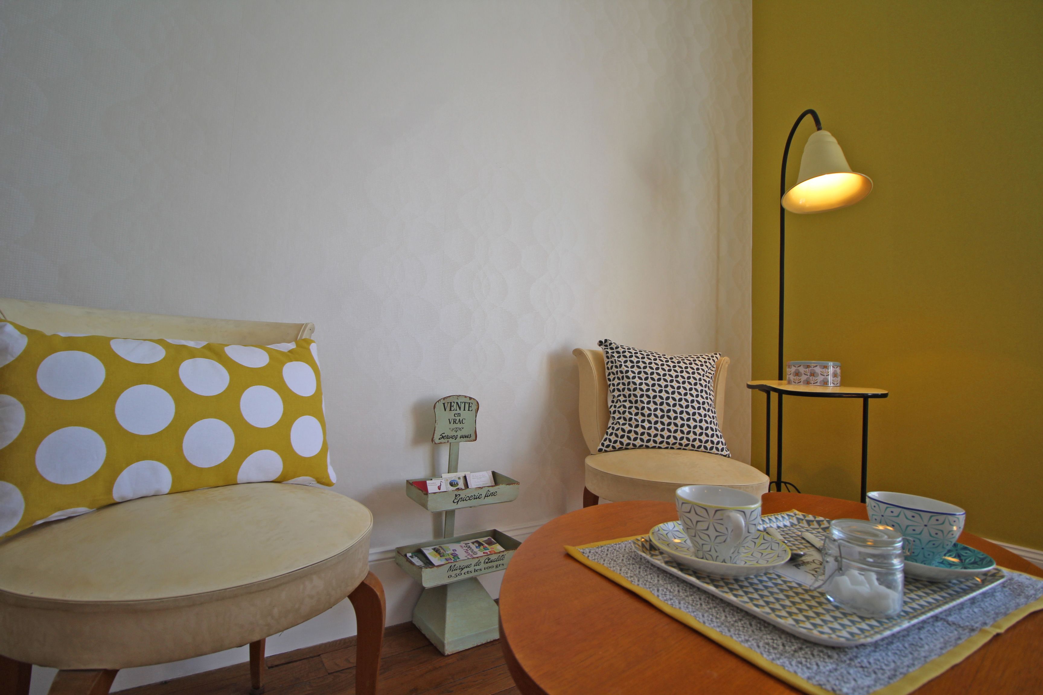 Una habitación acogedora con pared amarilla, suelo de madera, una mesa redonda con una bandeja de objetos, una silla con cojín de lunares y una lámpara sobre una mesita.