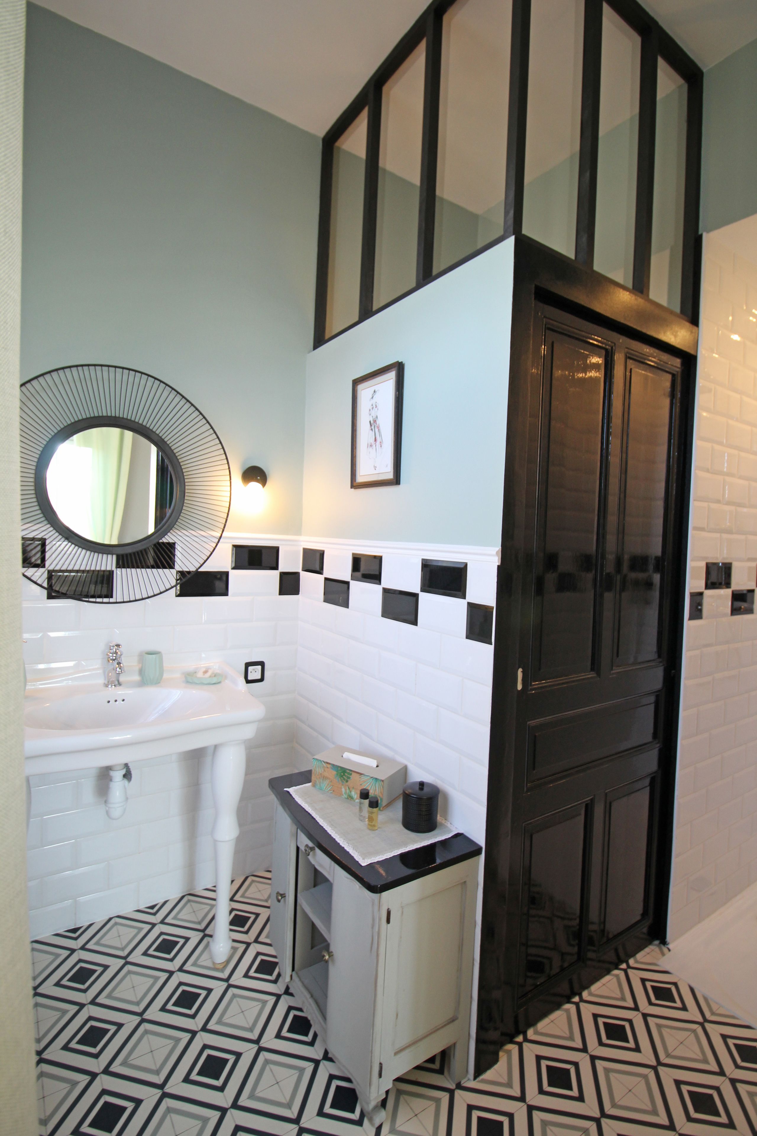 Un baño con un lavamanos blanco, un espejo, un mueble pequeño y una puerta negra.