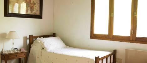 A small, cozy bedroom with a single bed, a nightstand, a framed picture on the wall, and a window with wooden shutters.
