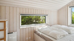 A bedroom with a wooden slatted ceiling, a window with a view of greenery, a bed with white bedding, and a wooden dresser.