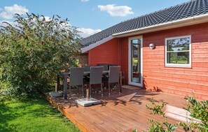 Una casa de madera roja con techo de tejas, un patio con mesa y sillas, y un jardín con diversas plantas.