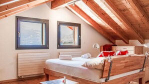 Une chambre avec un plafond en bois, deux fenêtres, un lit avec une literie blanche, une table de chevet et une commode.