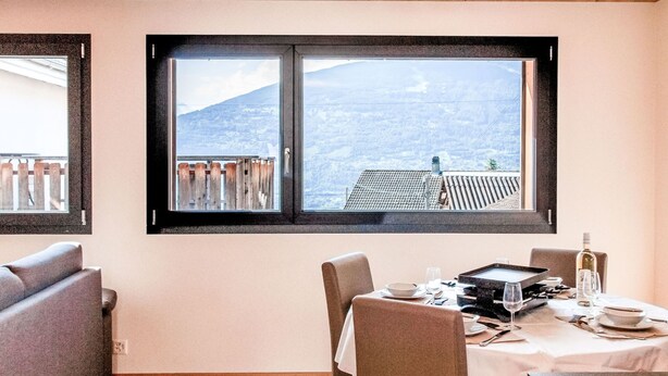 Un coin repas avec une table dressée pour deux personnes, une vue sur un paysage montagneux par une grande fenêtre et un plafond en bois.