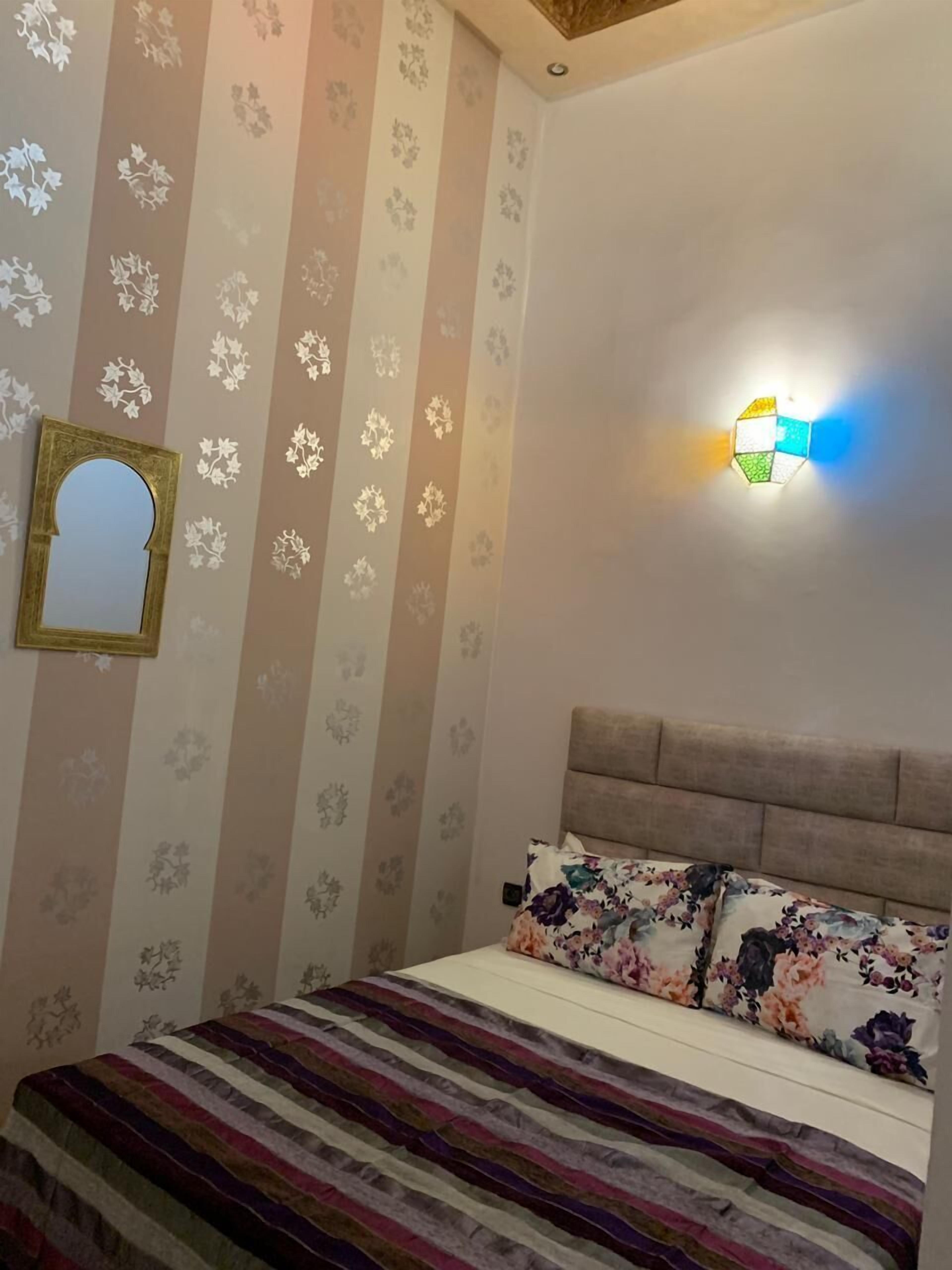 Une chambre avec un papier peint à rayures, un lit avec une literie à motifs floraux et un luminaire fixé au mur.