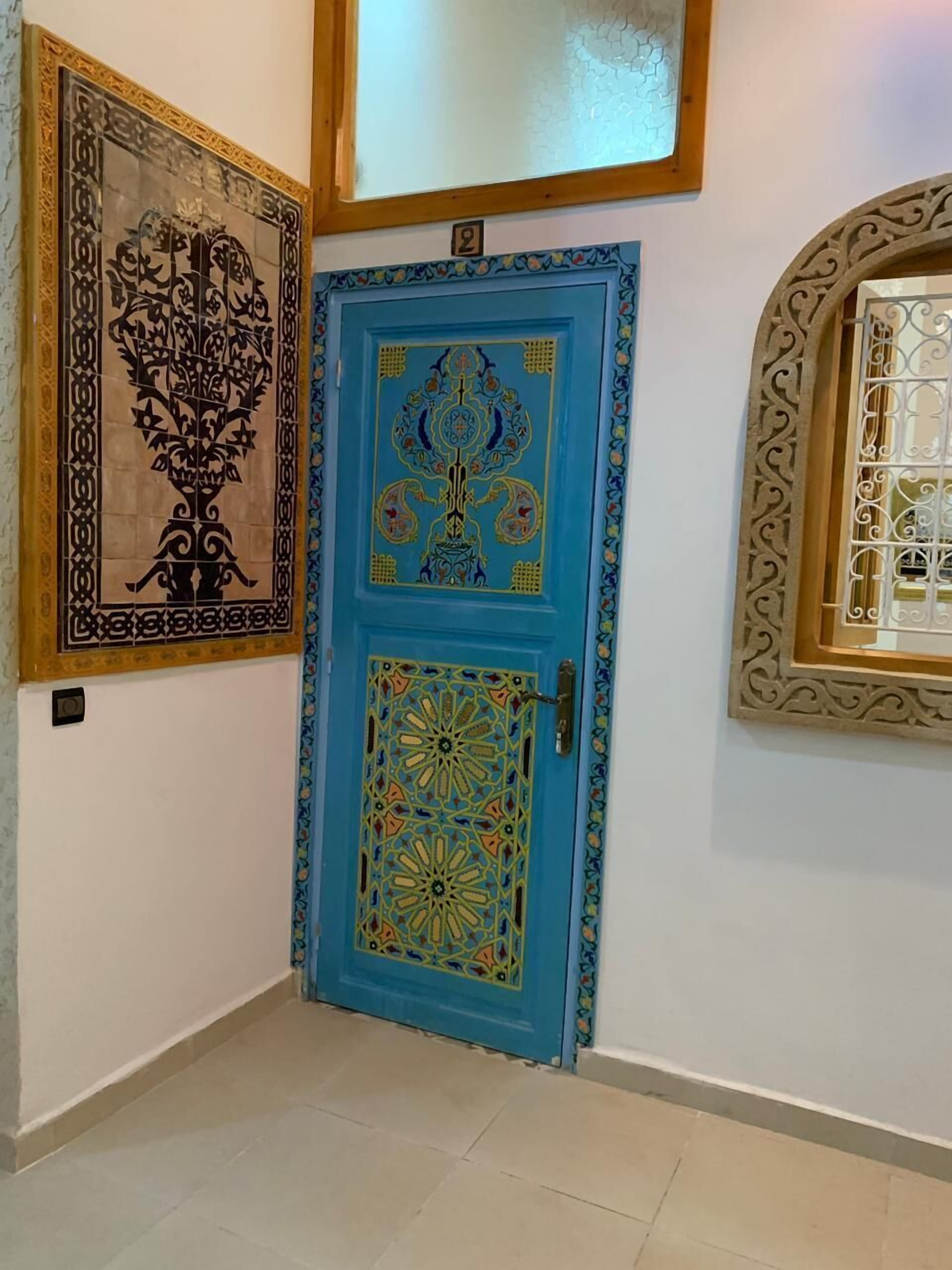 Un pasillo con una puerta azul con diseños intrincados, una obra de arte enmarcada en la pared y un ventanal decorativo.