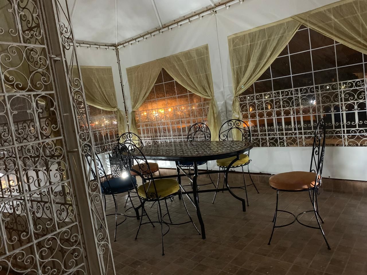 Un área de comedor al aire libre techada con sillas y una mesa de metal, rodeada por barandales de metal decorativos y cortinas.