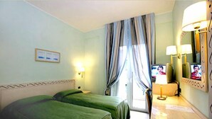 Ein Hotelzimmer mit zwei Einzelbetten, einem Schreibtisch mit Lampe, einem Fernseher und einem Fenster mit Vorhängen.