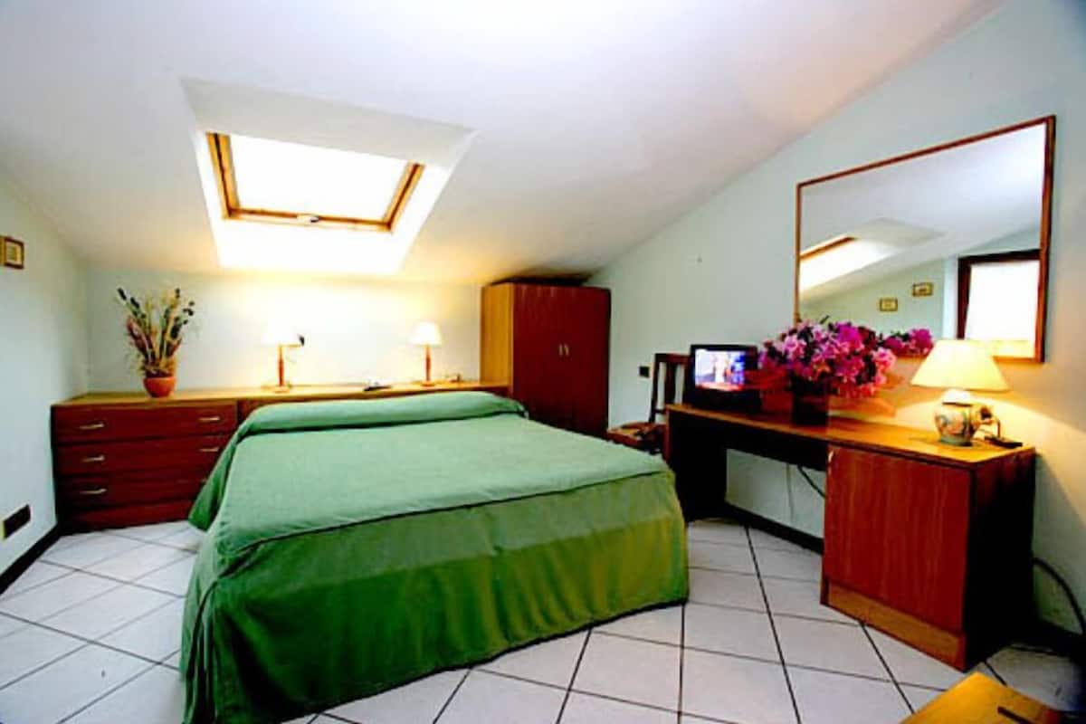 A hotel room with a bed, a desk with a computer, a mirror, and a window with a skylight.