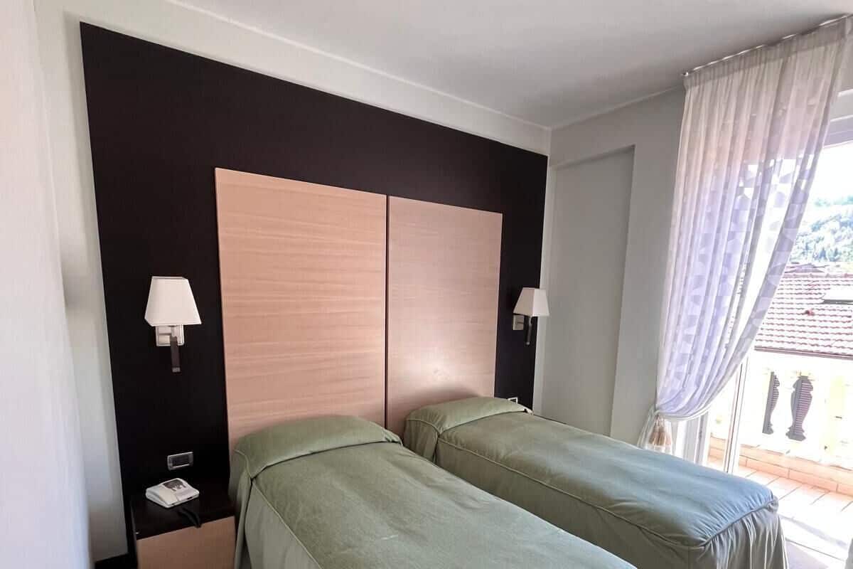 A hotel room with two single beds, a dark-colored headboard, bedside lamps, a small table with a phone, and a window with curtains.