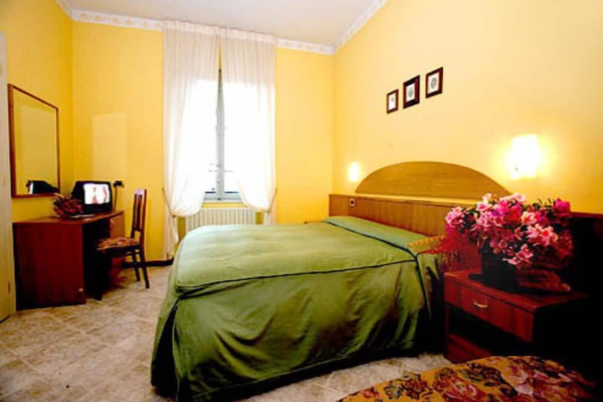 A hotel room with a green bed, a wooden headboard, a nightstand with a vase of flowers, a chair, a desk with a computer, and a window with white curtains.