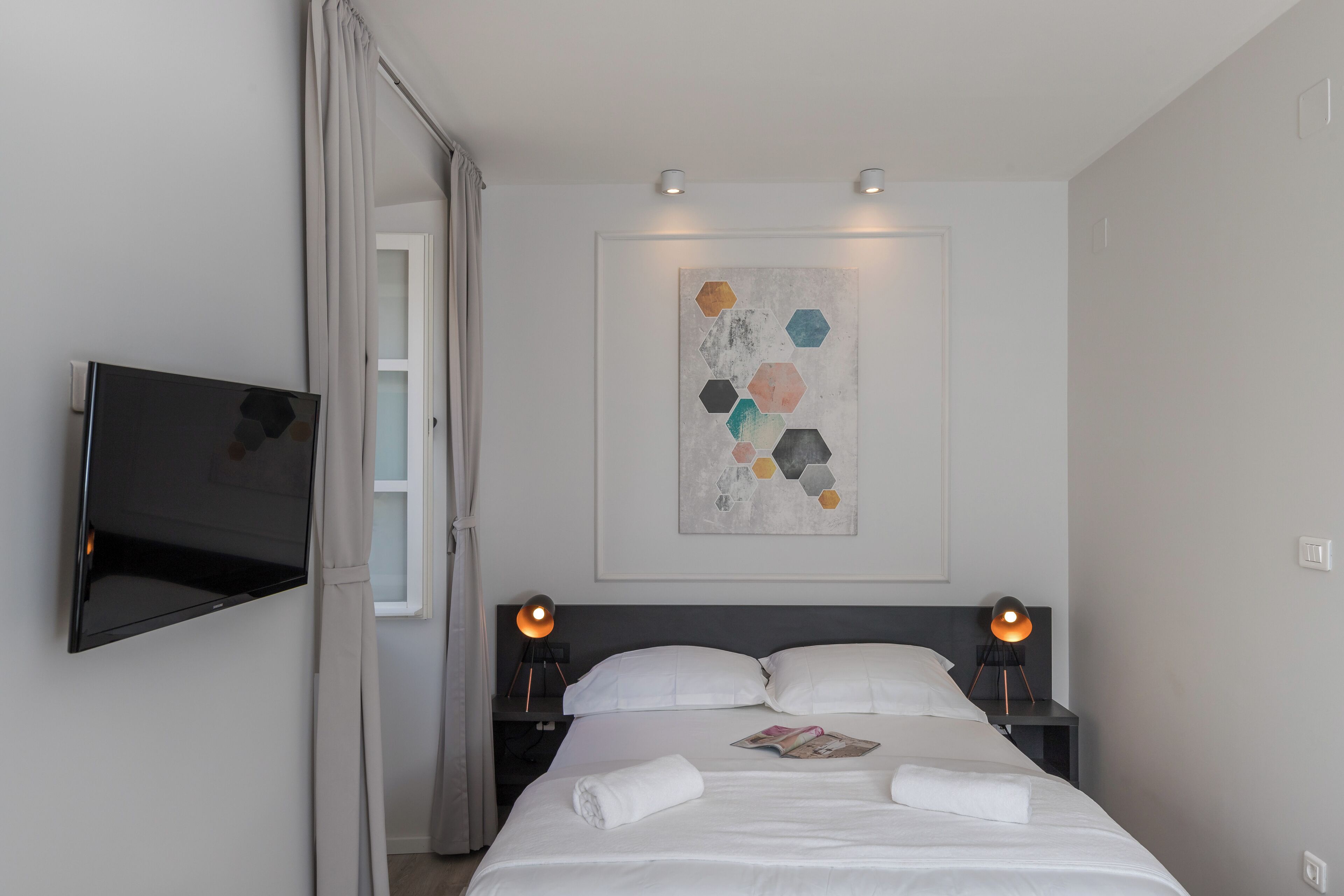 Una habitación de hotel moderna con una televisión de pantalla plana montada en la pared, una cama con ropa de cama blanca, dos lámparas de noche y un cuadro abstracto geométrico sobre la cama.