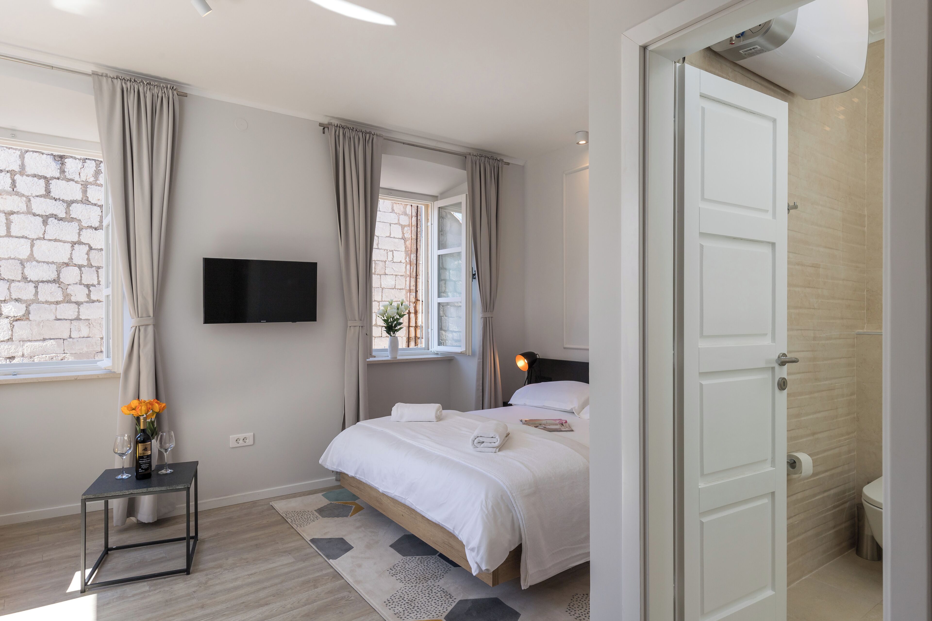 A modern bedroom with a bed, a TV mounted on the wall, a small table with a bottle and glasses, and a window with curtains.